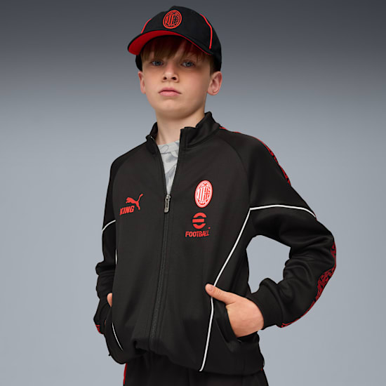 Vor grauem Hintergrund präsentiert ein Kind eine schwarze AC Milan Jacke und Kappe mit roten Akzenten.