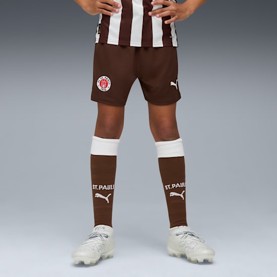 Op grijs draagt een model een St. Pauli-tenue: bruine shorts, sokken en witte hardloopschoenen.
