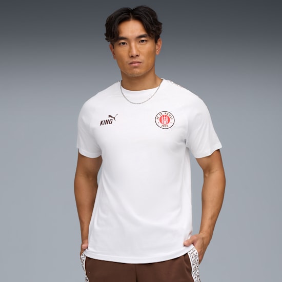 Een model poseert tegen een grijze achtergrond, draagt een wit t-shirt, bruine broek en ketting.