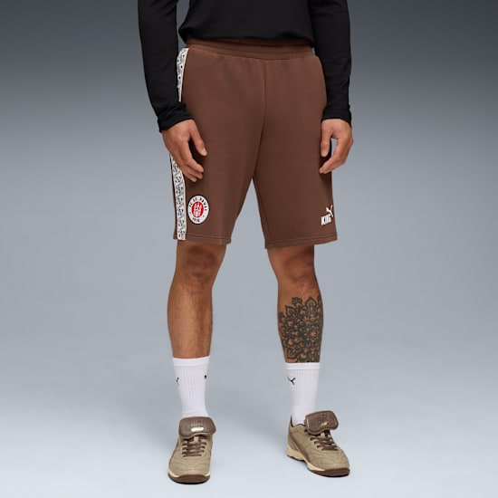Against grey, a model wears brown shorts, a black top, tube socks and trainers.
