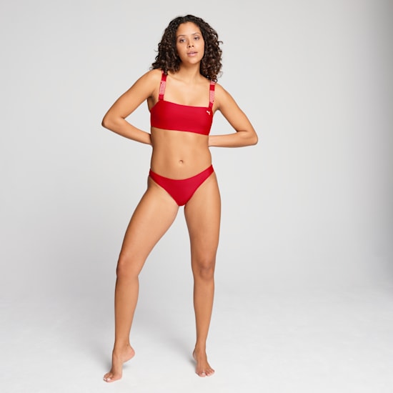 A model poses against a grey backdrop, wearing a red two-piece swimsuit with branded straps.