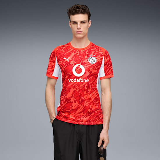 A model poses against a grey backdrop, wearing a red patterned t-shirt and black shorts.