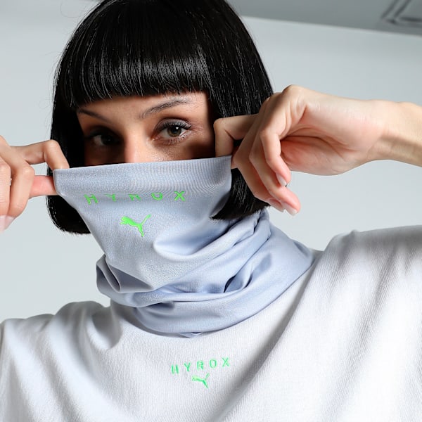 On a grey background, a model adjusts a light blue neck warmer with green logos.