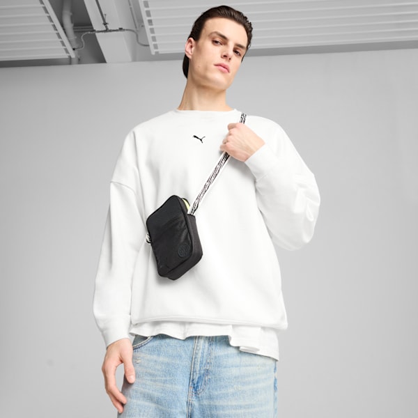 Against a grey backdrop, a model poses with a black cross-body bag and white jumper.