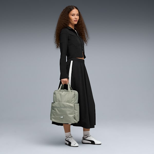 A model holds a green rucksack against a grey backdrop, wearing a black top and skirt with white trainers.