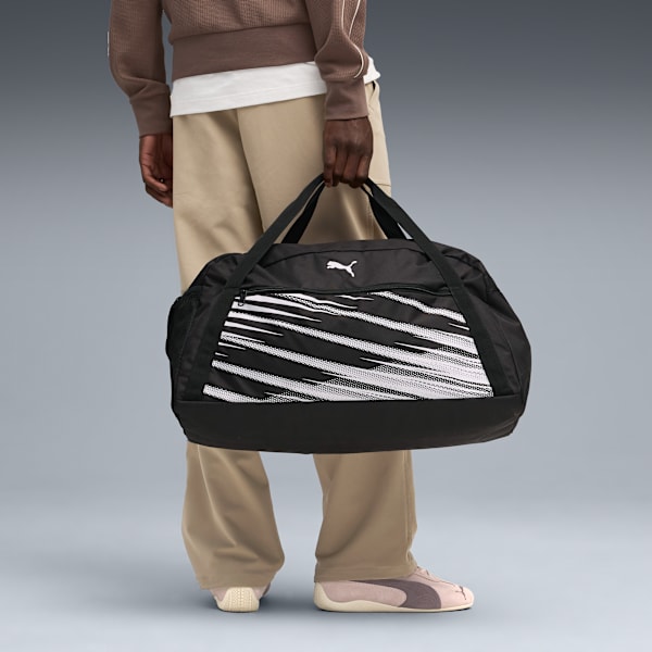 Against grey, a model holds a black duffel bag with white stripes and the Puma logo.