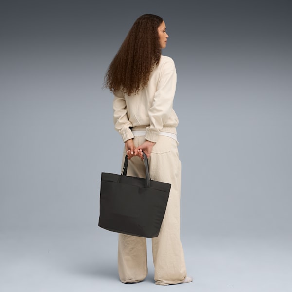 On grey, a model holds a black tote bag, wearing a beige jacket and trousers, looking to the side.