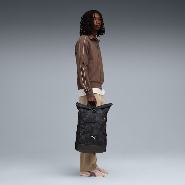 On grey, a model holds a dark patterned roll-top bag wearing brown jacket and trousers.