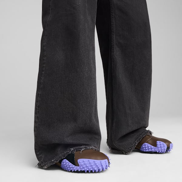 Against grey, a model wears wide black jeans and brown shoes with a textured, purple sole.