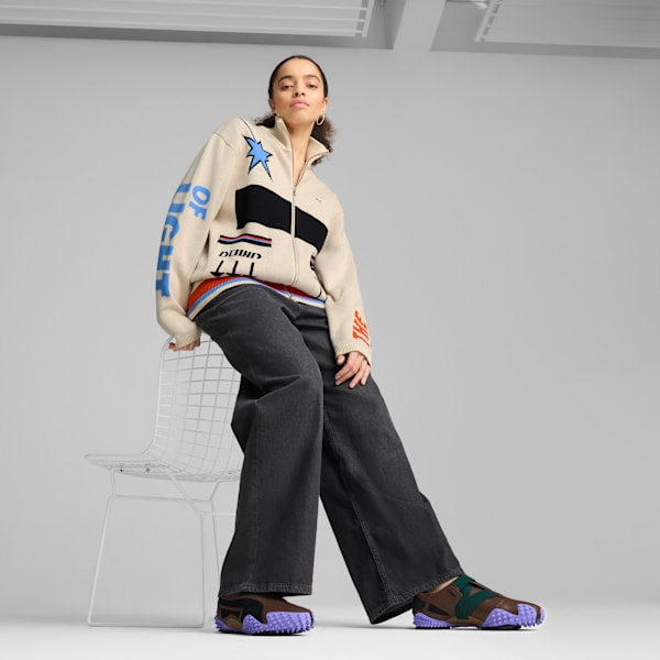 On grey, a model sits wearing patterned zip-up jacket, dark jeans and brown trainers.