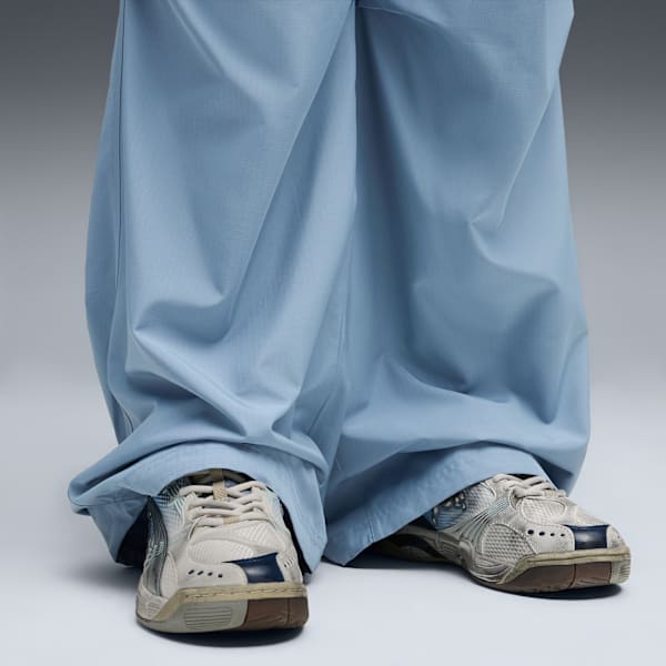 On grey, a model wears light blue trousers and neutral trainers with navy details.