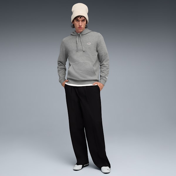 A model poses against a grey backdrop, wearing a grey hoodie, black trousers and a cream hat.