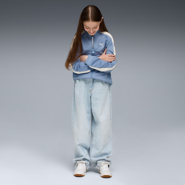 A model with crossed arms looks down, wearing a blue jacket, jeans and trainers on grey.