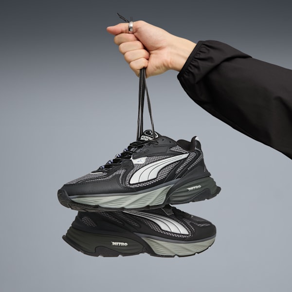 Against grey, a hand holds black Puma trainers by their laces, one shoe above its reflection.