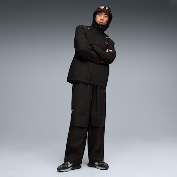 Against grey, a model in dark hooded jacket and trousers stands with arms crossed.