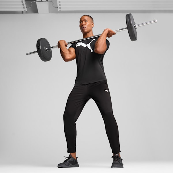Against grey, a model lifts a barbell wearing a black Puma t-shirt and black joggers.