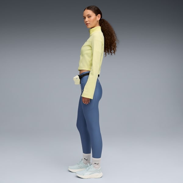 A model poses against grey wearing a yellow top, blue leggings, trainers and a waist bag.