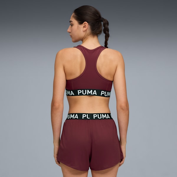 A model faces left against grey, wearing a maroon sports bra and shorts with logo detail.