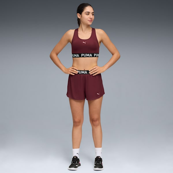 Against a grey backdrop, a model poses in a maroon sports bra, shorts, and running shoes.