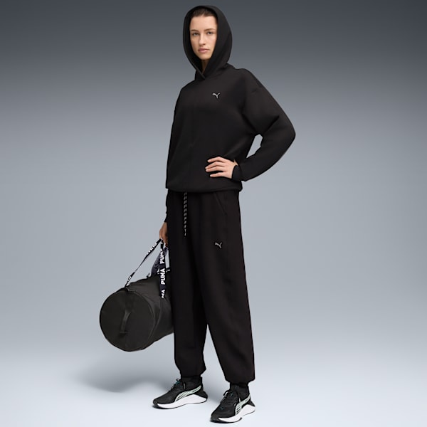 A model poses against grey backdrop, wearing black Puma hooded tracksuit and carrying a bag.