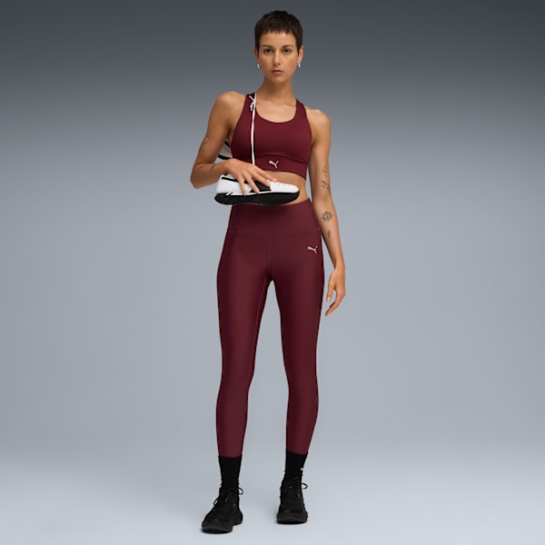 A model against grey holds a shoe, wearing a maroon sports bra and leggings.