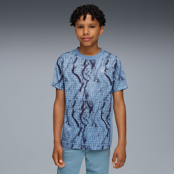 A model with curly hair stands against a grey backdrop, wearing a patterned blue t-shirt and blue bottoms.