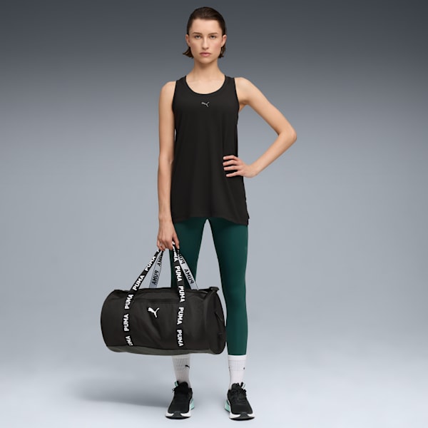 Model poses with a branded holdall, wearing a black top, green leggings and trainers against grey.