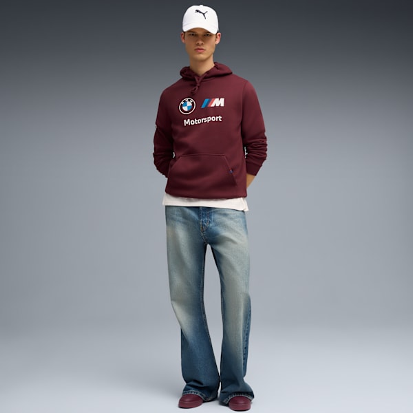 A model stands against grey, wearing a branded hoodie, jeans, and a white cap.