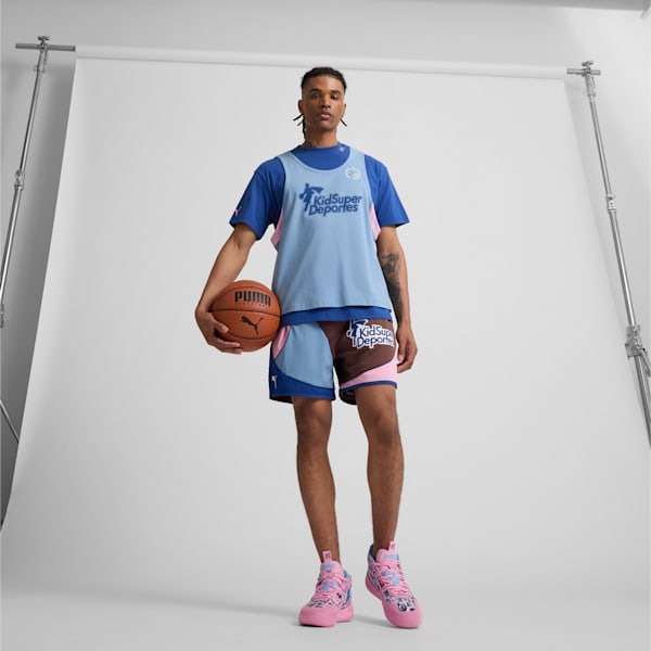 On a white backdrop, a model holds a basketball, wearing a blue t-shirt, shorts and pink trainers.