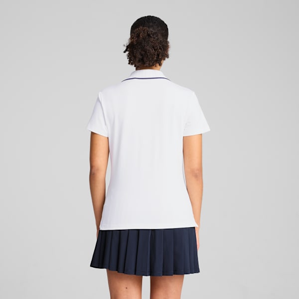 A model faces away on grey, wearing a white polo shirt and a short, dark blue pleated skirt.