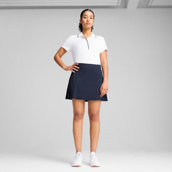 Against a grey backdrop, a model poses wearing a white polo shirt, a navy skirt and white trainers.
