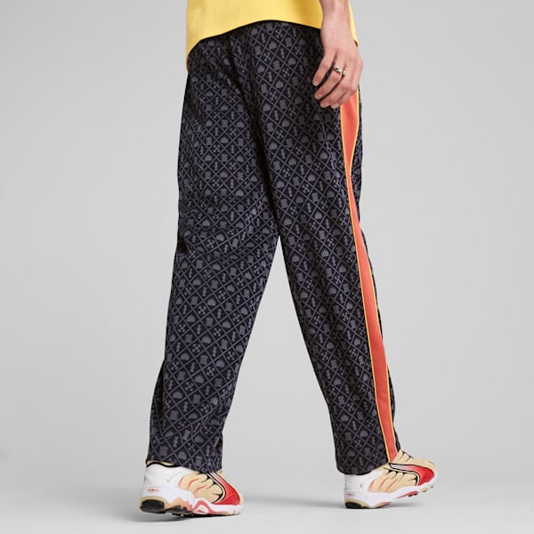 Against grey, a model wears patterned black trousers with a red stripe and colourful trainers.