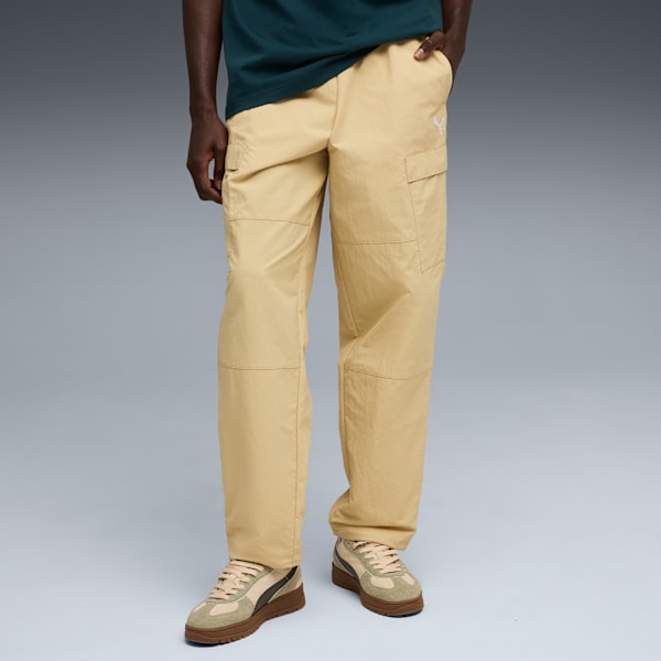 On grey, a model wears beige trousers, a dark green t-shirt, and beige-and-brown trainers.