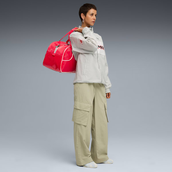 A model stands with a red bag on shoulder against a grey backdrop, wearing a jacket and trousers.