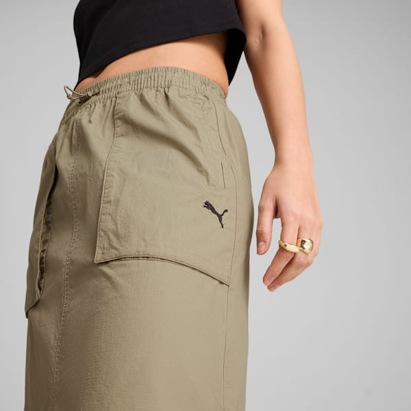 Against grey, a model wears olive trousers with a black logo and a gold ring.