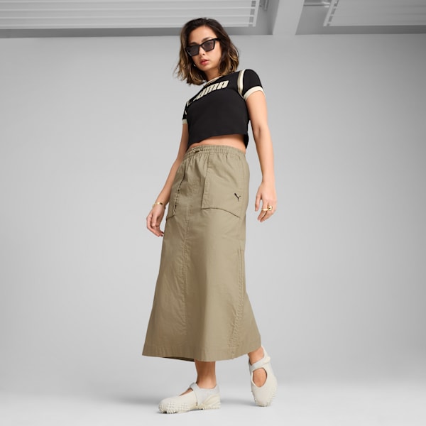 A model poses against a grey backdrop, wearing a black top, khaki skirt, and light shoes.