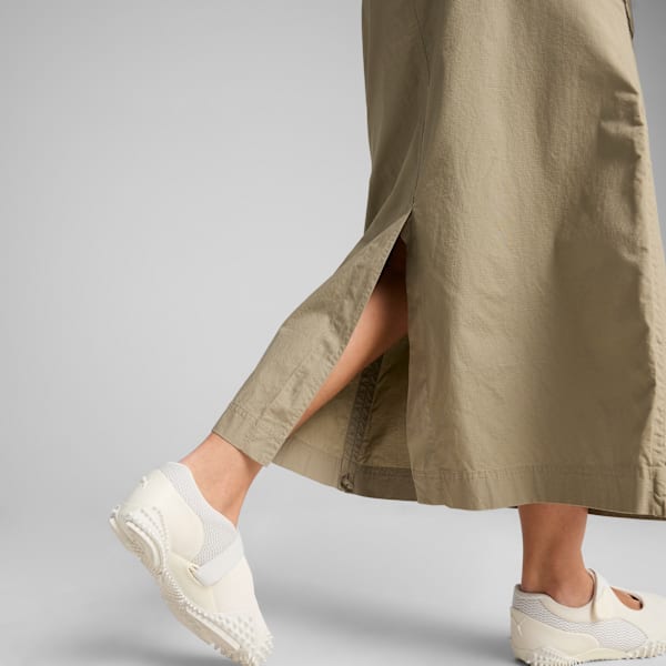 Against grey, a model moves in a long olive skirt and off-white shoes.