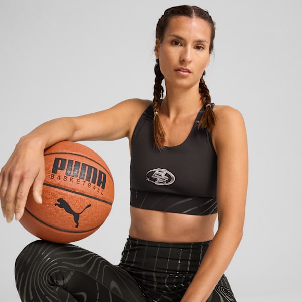 A model with braids poses with a basketball against a grey backdrop, wearing a sports bra.