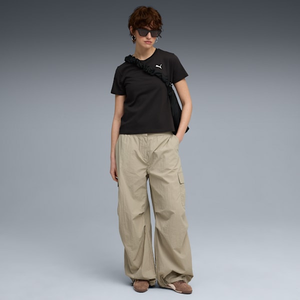 On a grey backdrop, a model poses in a black tee, beige pants, and trainers.