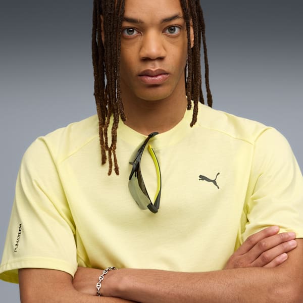 Against grey, a model with dreadlocks wears a yellow t-shirt and sunglasses.
