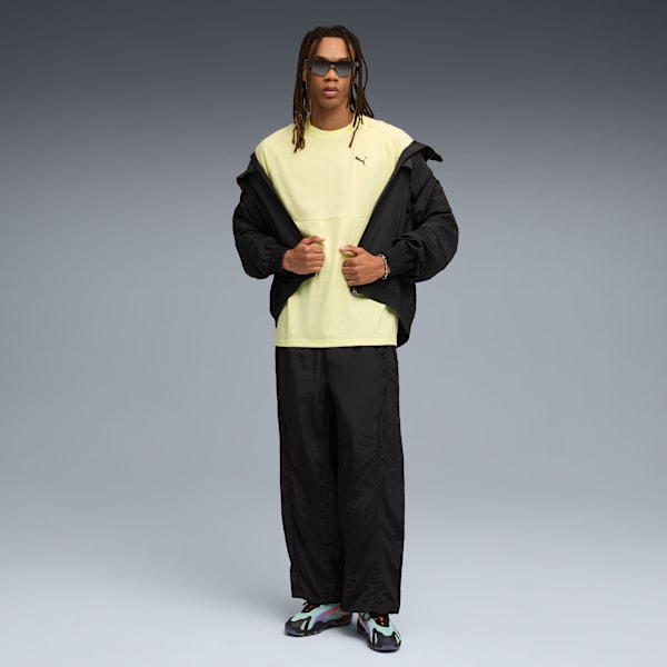 On grey, a model poses in a yellow t-shirt, black jacket, trousers, and trainers.
