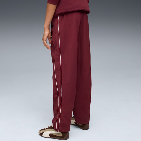 Against grey, a model wears maroon trousers with a white stripe and brown trainers.