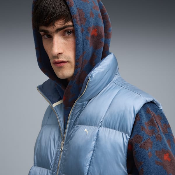 Against a grey backdrop, a model poses wearing a blue gilet over a patterned hooded top.