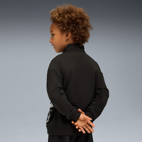 A child poses against grey wearing a black zip-up top with hands clasped behind.