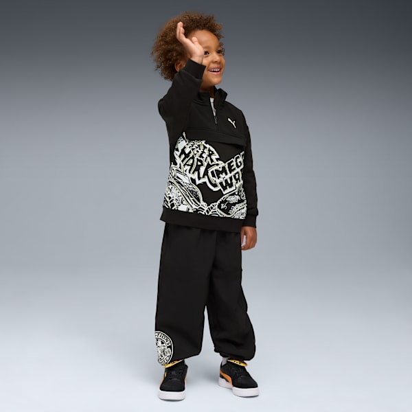Against grey, a child smiles, hand raised, wearing a black graphic sweatshirt and trousers.