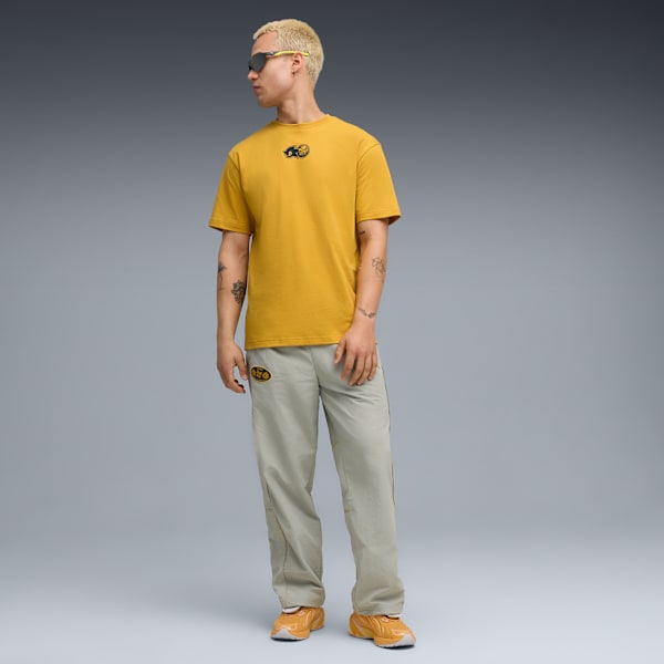 Against grey, a model poses in a gold t-shirt, light trousers and orange trainers.