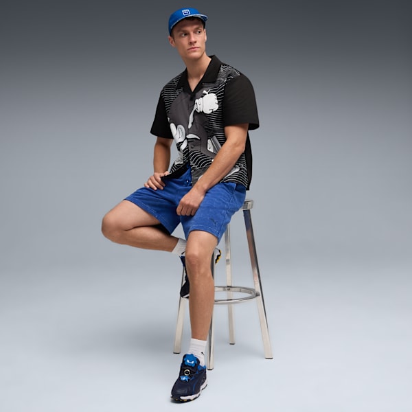 A model sits on a stool against a grey backdrop, wearing a patterned shirt and blue shorts.