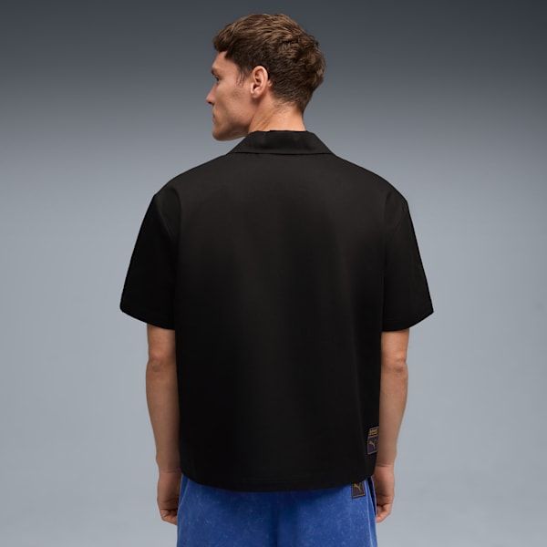 A model faces away on grey, wearing a black shirt and blue shorts while looking left.