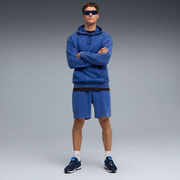A model with crossed arms wears a blue hoodie and shorts against a grey backdrop.