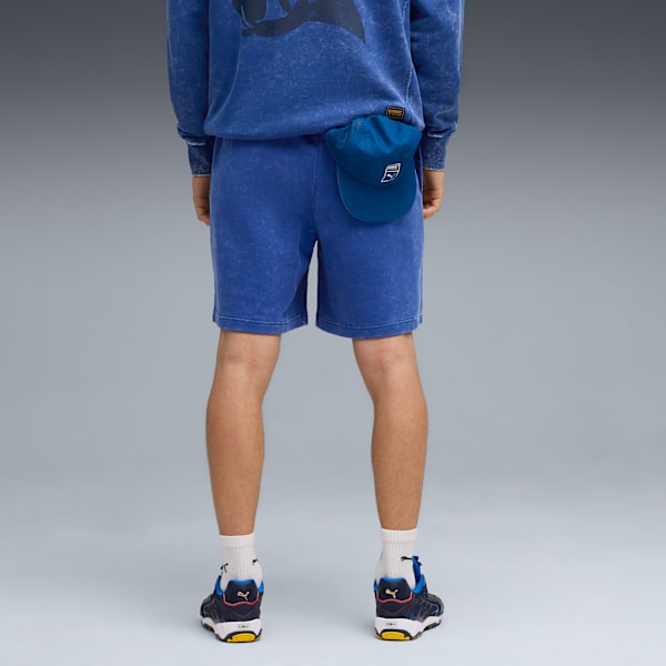 Model stands against grey backdrop, wearing blue shorts, top, trainers and a cap.